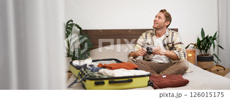 Portrait of happy, smiling young man sitting on bed, unpacking his suitcase after a holiday, tourist looking at his photo album on camera 126015175