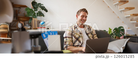Portrait of young handsome man sitting with laptop, has passport and two plane tickets prepared, planning his journey on computer, booking hotels and restaurants online for the trip 126015285