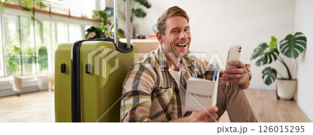 Portrait of smiling handsome tourist, man holding passport and flight tickets, sitting with suitcase Portrait of smiling handsome tourist, man holding passport and flight tickets, sitting with suitcase 126015295