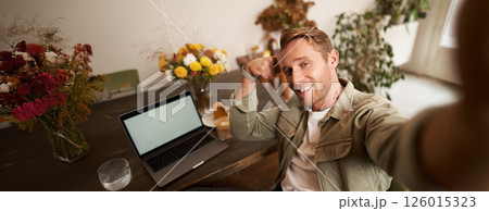 Portrait of handsome and confident man taking a selfie, flexing biceps, shows-off his muscles, sits in cafe in front of laptop, smiling at camera 126015323