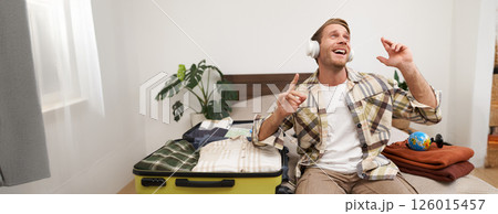 Portrait of cheerful handsome man, tourist going on vacation, dancing and listening to music in headphones while packing suitcase with clothes for holiday trip abroad 126015457