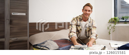 Portrait of young man, tourist going travelling abroad, putting clothes in suitcase, closing his luggage, sitting on bed, getting ready for holiday 126015581