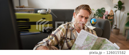 Portrait of happy young man, tourist with suitcase, sits in living room and looks at map, chooses holiday destination, picks route for travelling Portrait of happy young man, tourist with suitcase, sits in living room and looks at map, chooses holiday destination, picks route for travelling 126015582