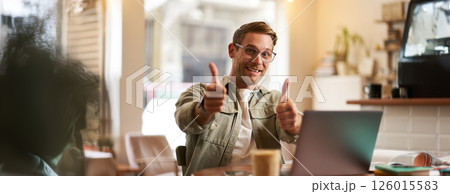 Joyful smiling guy in glasses, man shows thumbs up, sits in cafe with laptop, recommends website, e-learning service or co-working space Joyful smiling guy in glasses, man shows thumbs up, sits in cafe with laptop, recommends website, e-learning service or co-working space 126015583