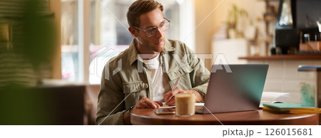 Portrait of handsome young guy in glasses, man studying, taking notes, looking at laptop screen during online lesson, attends a meeting and writing down information, sitting in cafe Portrait of handsome young guy in glasses, man studying, taking notes, looking at laptop screen during online lesson, attends a meeting and writing down information, sitting in cafe 126015681