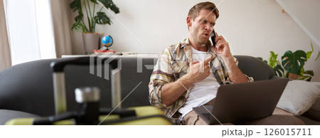 Portrait of angry man arguing over the phone, has laptop opened on his laptop, sits near suitcase, complaining at travel agency 126015711
