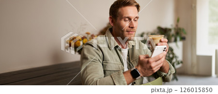 Lifestyle portrait of handsome young man, sitting in cafe, checking his phone and drinking coffee, smiling. 126015850