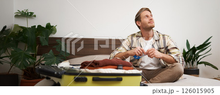 Portrait of handsome man, tourist, looking at his holiday tickets, booked vacation with travel agency, sitting on bed and packing clothes in suitcase Portrait of handsome man, tourist, looking at his holiday tickets, booked vacation with travel agency, sitting on bed and packing clothes in suitcase 126015905