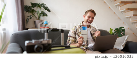 Excited young man, looking happy at laptop, sitting with flight tickets and passport, has suitcase packed for holiday trip 126016033