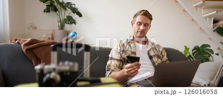 Portrait of cheerful guy buying flight tickets online, booking hotel by using laptop and credit card, sitting near suitcase in living room Portrait of cheerful guy buying flight tickets online, booking hotel by using laptop and credit card, sitting near suitcase in living room 126016058