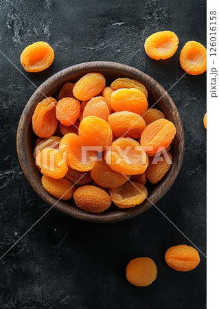 A wooden bowl brimming with dried apricots, some scattered around, their vibrant orange color contrasting beautifully with the dark, textured surface below. 126016158