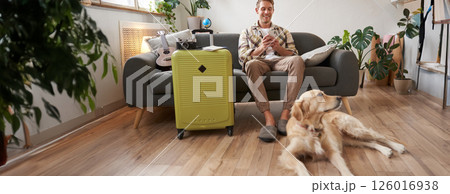 Portrait of happy, handsome young man with suitcase, sitting with his dog in living room, prepared to go on vacation, going on holiday Portrait of happy, handsome young man with suitcase, sitting with his dog in living room, prepared to go on vacation, going on holiday 126016938