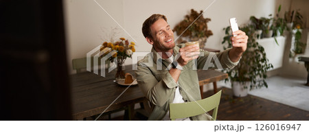 Portrait of young handsome man takes slefie on his mobile phone app, sits in cafe with glass of coffee, posting on social media application Portrait of young handsome man takes slefie on his mobile phone app, sits in cafe with glass of coffee, posting on social media application 126016947