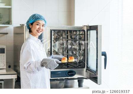 オーブンでパンを焼く衛生白衣を着た女性調理師 給食センター 給食室 オーブンでパンを焼く衛生白衣を着た女性調理師 給食センター 給食室 126019337