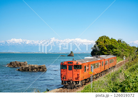 富山湾に浮かぶ男岩と残雪の立山連峰を背景に雨晴海岸を走るJR氷見線の気動車 126019372