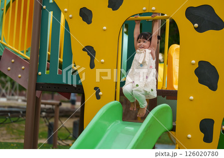 happy toddler girl hanging the yellow bar by her hand at outdoor playground in park 126020780
