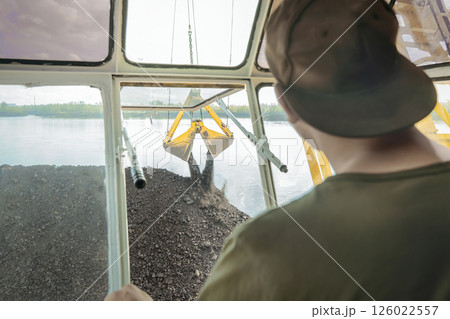 Loading coal onto a barge ship on a floating crane, view from the crane operator's cabin, rear view of the crane operator. A crane pours coal onto a barge. Transportation of coal by sea or river. Loading coal onto a barge ship on a floating crane, view from the crane operator's cabin, rear view of the crane operator. A crane pours coal onto a barge. Transportation of coal by sea or river. 126022557