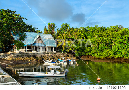 Small fishing boats are moored along a quiet shore near a tin-roofed coastal house in Koror, Palau, framed by calm waters and lush tropical greenery in the early morning light. Small fishing boats are moored along a quiet shore near a tin-roofed coastal house in Koror, Palau, framed by calm waters and lush tropical greenery in the early morning light. 126025403