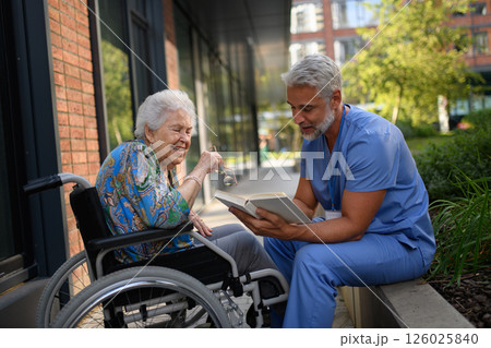 Male caregiver reading book to senior woman in wheelchair. 126025840