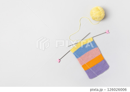 Striped piece of knitted fabric for future clothing with knitting needles and yellow ball of thread on gray background. Homemade needlework and knitting of warm, clothes and toys. Concept creativity 126026006