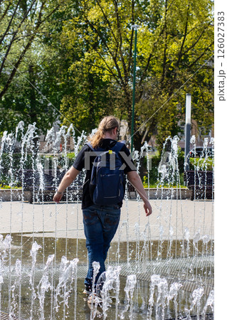 A man carrying a backpack is leisurely walking through a beautiful fountain A man carrying a backpack is leisurely walking through a beautiful fountain 126027383
