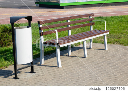 There is a wooden bench situated alongside a white trash can on a sidewalk 126027506
