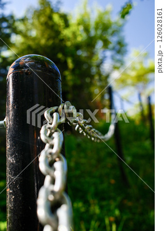 Black metal park bollard with attached security chain, close-up of boundary post with galvanized steel chain links 126028161