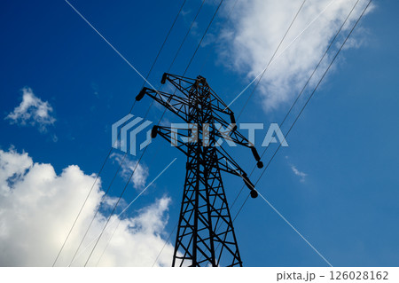 Electrical transmission tower silhouette against bright blue sky with scattered white clouds, high voltage power line pylon 126028162