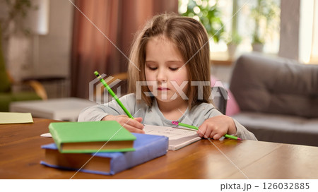 Child girl sitting at table, writing in notebook with pencils in living room at home. Beginning of learning, preschool development Child girl sitting at table, writing in notebook with pencils in living room at home. Beginning of learning, preschool development 126032885