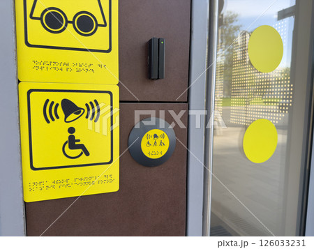 Accessibility call system with yellow tactile and audio symbols, including Braille text and assistance button near glass door, designed to support people with disabilities in modern infrastructure 126033231