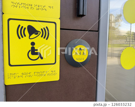 Yellow tactile signage and assistance button with bell and disability symbols including Braille, providing accessible user interface near modern entrance for inclusive and barrier free access Yellow tactile signage and assistance button with bell and disability symbols including Braille, providing accessible user interface near modern entrance for inclusive and barrier free access 126033232