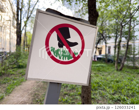 Warning sign in urban park prohibiting walking on the grass. Signpost with red circle and black boot pictogram placed in a green public area surrounded by trees and buildings 126033241