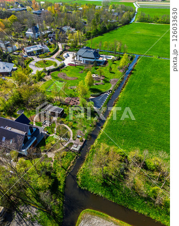 Aerial view of a modern countryside neighborhood with green lawns, small canals, landscaped gardens, and residential houses surrounded by fields and patches of forest. 126033630