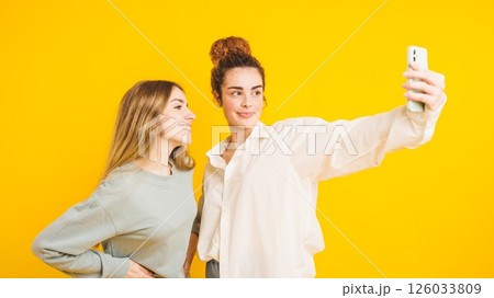 Two young women taking a selfie with smartphone on yellow background Two young women taking a selfie with smartphone on yellow background 126033809