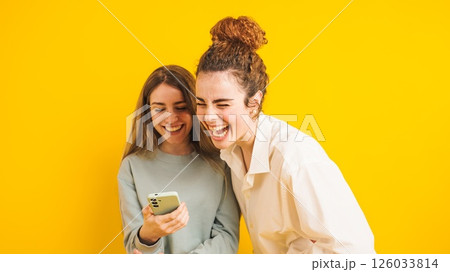 Two young women laughing while looking at smartphone on yellow background 126033814