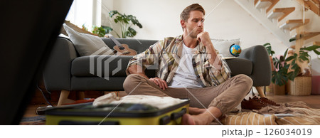 Portrait of stressed young man packing his suitcase, deciding what clothes bring on vacation, thinking while looking at luggage Portrait of stressed young man packing his suitcase, deciding what clothes bring on vacation, thinking while looking at luggage 126034019