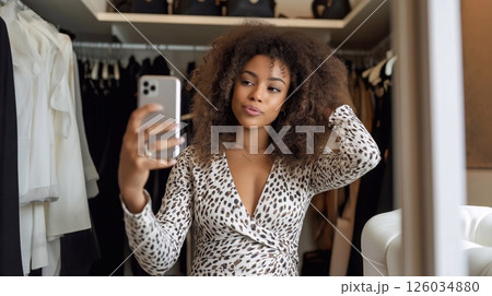 Fashion blogger taking selfie in stylish wardrobe, showing trendy outfit and curly hair, creating content for social media and online shopping inspiration. 126034880