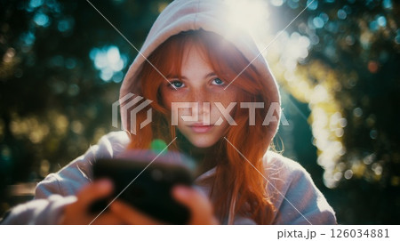 Teen girl with red hair and freckles in hoodie using smartphone outdoors. Concept of youth, gadget addiction, online safety, and digital generation. 126034881