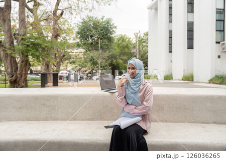 Muslim businesswoman sitting outdoors with a laptop and notebook, smiling. Muslim businesswoman sitting outdoors with a laptop and notebook, smiling. 126036265