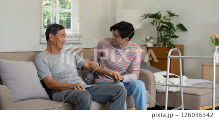 Joyful son sharing a moment with elderly father on the couch, highlighting family bonds and support in a cozy living room. 126036342