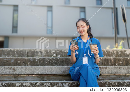 Nurse sitting on steps, smiling while using her smartphone and holding a coffee cup in a sunny healthcare environment Nurse sitting on steps, smiling while using her smartphone and holding a coffee cup in a sunny healthcare environment 126036898