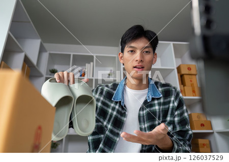 Young entrepreneur showcasing footwear during online presentation, holding shoes in modern workspace with product boxes. Young entrepreneur showcasing footwear during online presentation, holding shoes in modern workspace with product boxes. 126037529