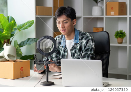 Young entrepreneur selling products online, using smartphone and laptop in modern workspace with greenery. Young entrepreneur selling products online, using smartphone and laptop in modern workspace with greenery. 126037530