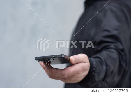 A man in a black jacket holding a smartphone against a gray concrete wall. Modern technology and the use of mobile devices. A man in a black jacket holding a smartphone against a gray concrete wall. Modern technology and the use of mobile devices. 126037766