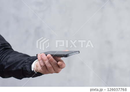 A man in a black jacket holding a smartphone against a gray concrete wall. Modern technology and the use of mobile devices. A man in a black jacket holding a smartphone against a gray concrete wall. Modern technology and the use of mobile devices. 126037768