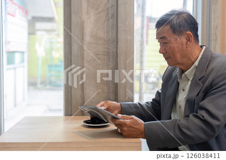 Senior businessman focused on a tablet while enjoying coffee in a modern cafe setting. Senior businessman focused on a tablet while enjoying coffee in a modern cafe setting. 126038411