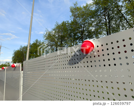 Close up of red warning beacons mounted on perforated metal fence. The setup suggests a security or restricted area, with modern infrastructure and safety signaling under clear sky 126039797