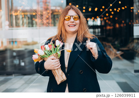 Beautiful young woman with spring tulips flowers bouquet walking at city street. Happy girl posing outdoors. Spring portrait of pretty female on urban background. High quality photo. 126039911