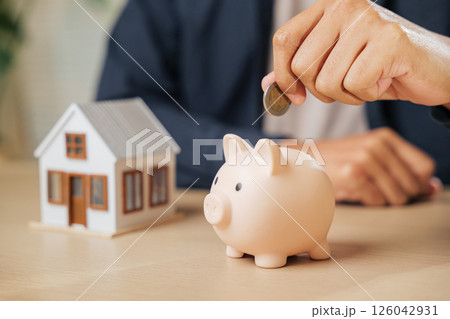 A businessman hand saving money by placing a coin into a piggy bank next to a house model, representing investment, financial stability, property goals, and a savings-focused approach to home buying. 126042931