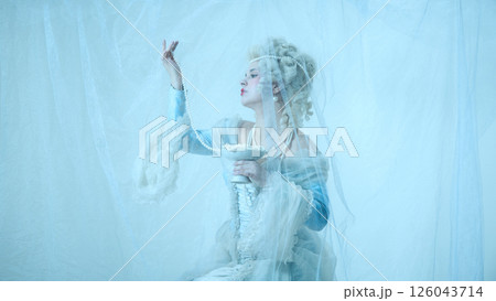 Young graceful woman as medieval duchess in vintage corset raising pearl necklace against light blue background. 126043714